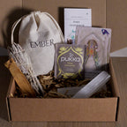 Open cardboard box on a wooden surface with various items including a white sage, palo santo and selenite.