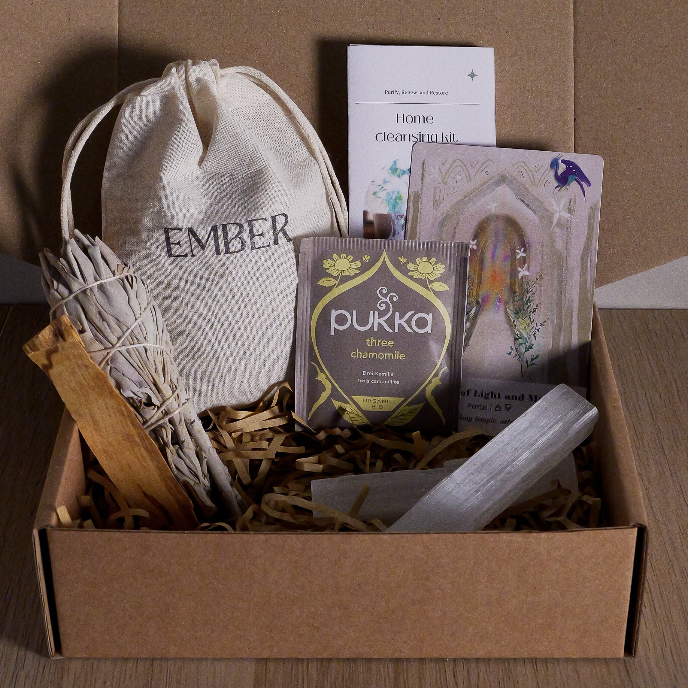 Open cardboard box on a wooden surface with various items including a white sage, palo santo and selenite.