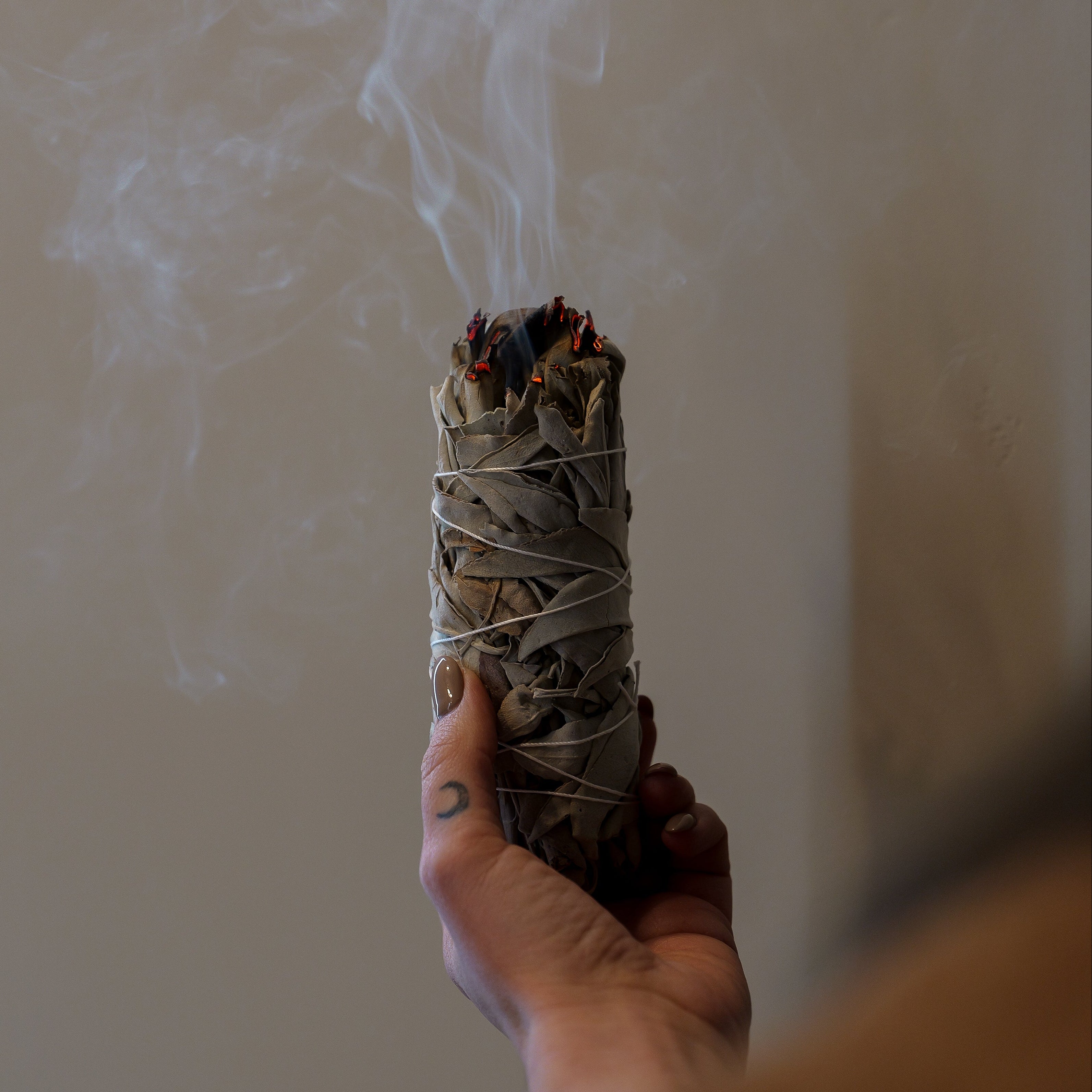 Hand holding a smoldering sage bundle with smoke against a neutral background