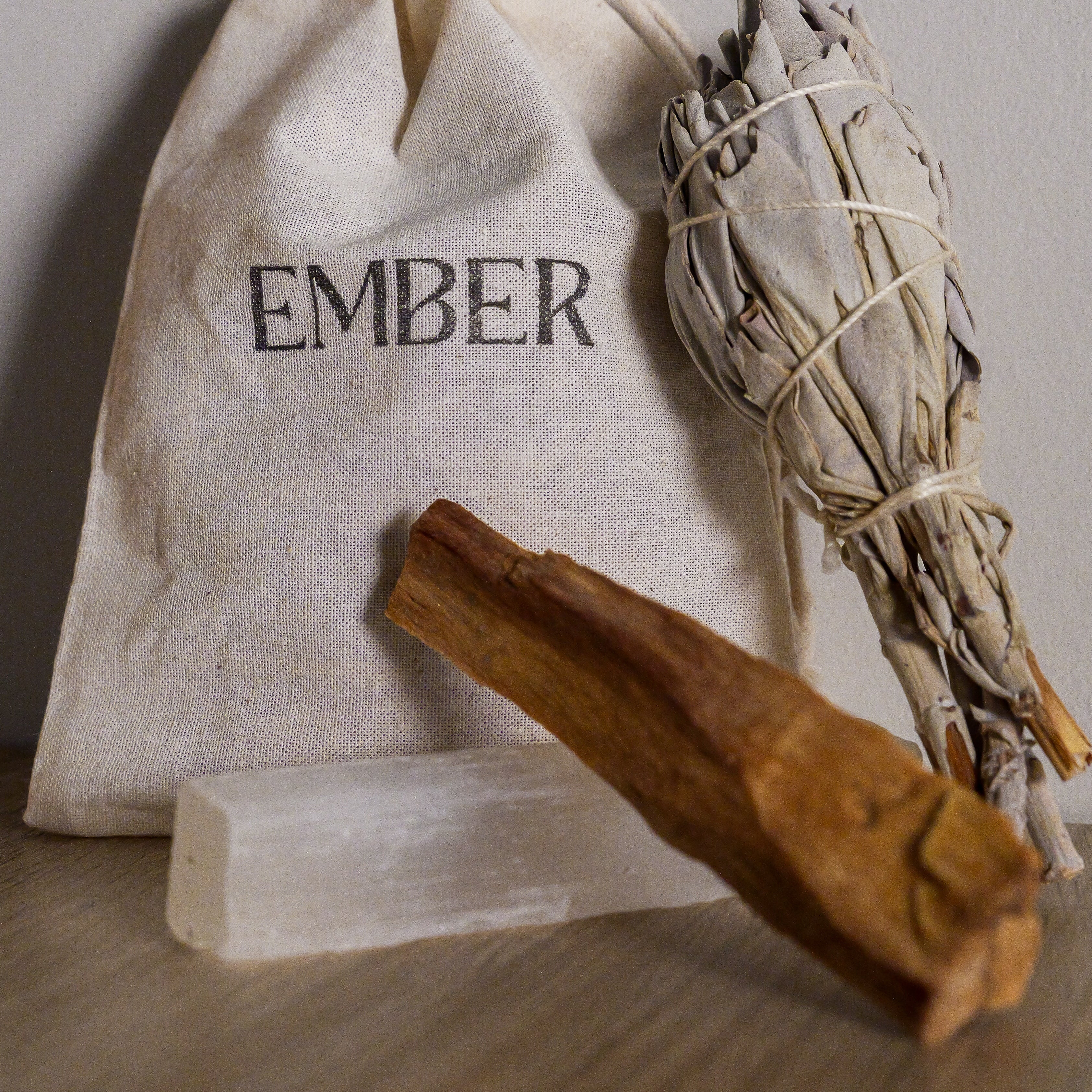White drawstring bag labeled 'EMBER' with a palo santo and dried herbs on a neutral background