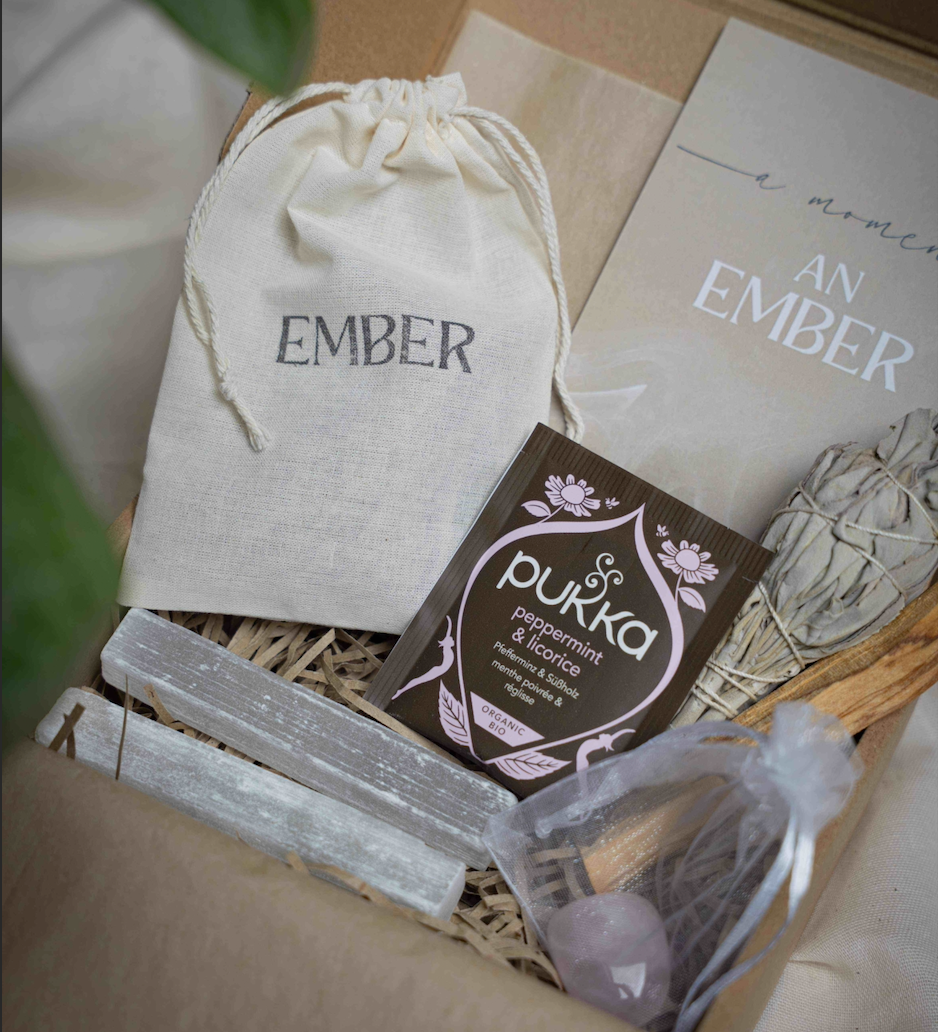 A curated gift box set featuring a cream drawstring pouch with 'EMBER' printed on it, a brown Pukka peppermint and lemon packet, white sage bundles, silver crystal points, and a clear bag with a pink crystal, arranged in a kraft paper box.