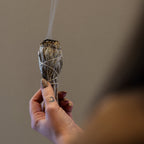 Hand holding a smoldering sage bundle with smoke against a neutral background