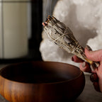 Hand holding a smoldering sage stick over a wooden bowl with a geode crystal in the background.